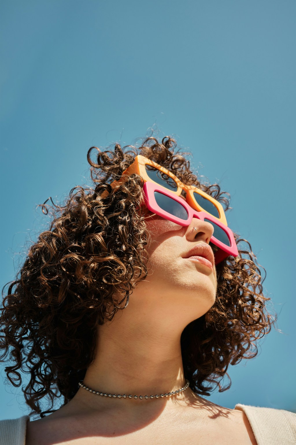 Image: a woman with curly brown hair looks up to the sun on a cloudless day, wearing chunky pink-framed sunglasses over her eyes, with another similar yellow-colored pair stacked right atop