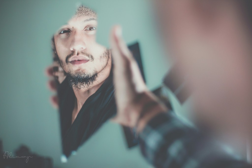 Image: a man with a short black beard and moustache stares at himself through a broken mirror shard - we see part of him from a blurred side-angle perspective, the only clear image of him through the shard