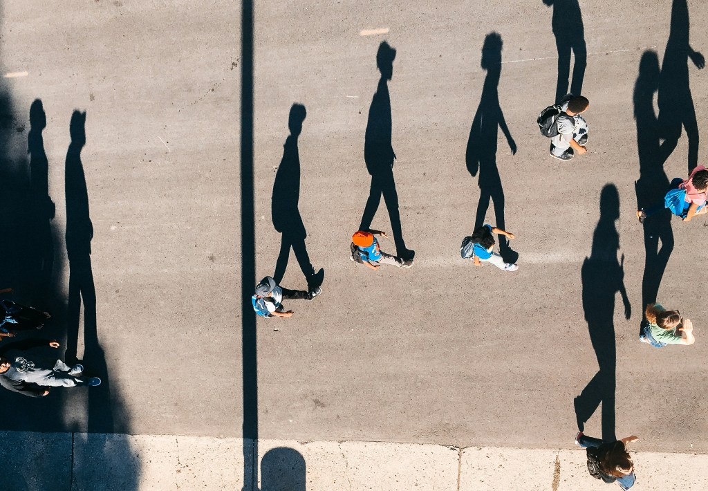 Image: bird's-eye shot of people walking on a street on a sunny day, their shadows exaggerated as if they were their own people
